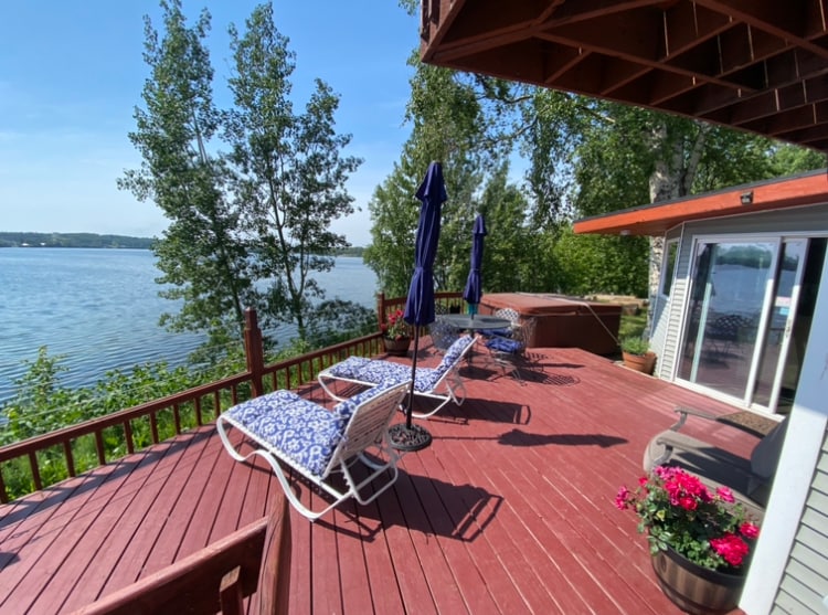 A waterfront deck offers multiple lounge chairs and vibrant umbrellas, providing a relaxing space to enjoy the lake views. Potted flowers add a touch of color, while the calm water reflects the surrounding trees and sky.