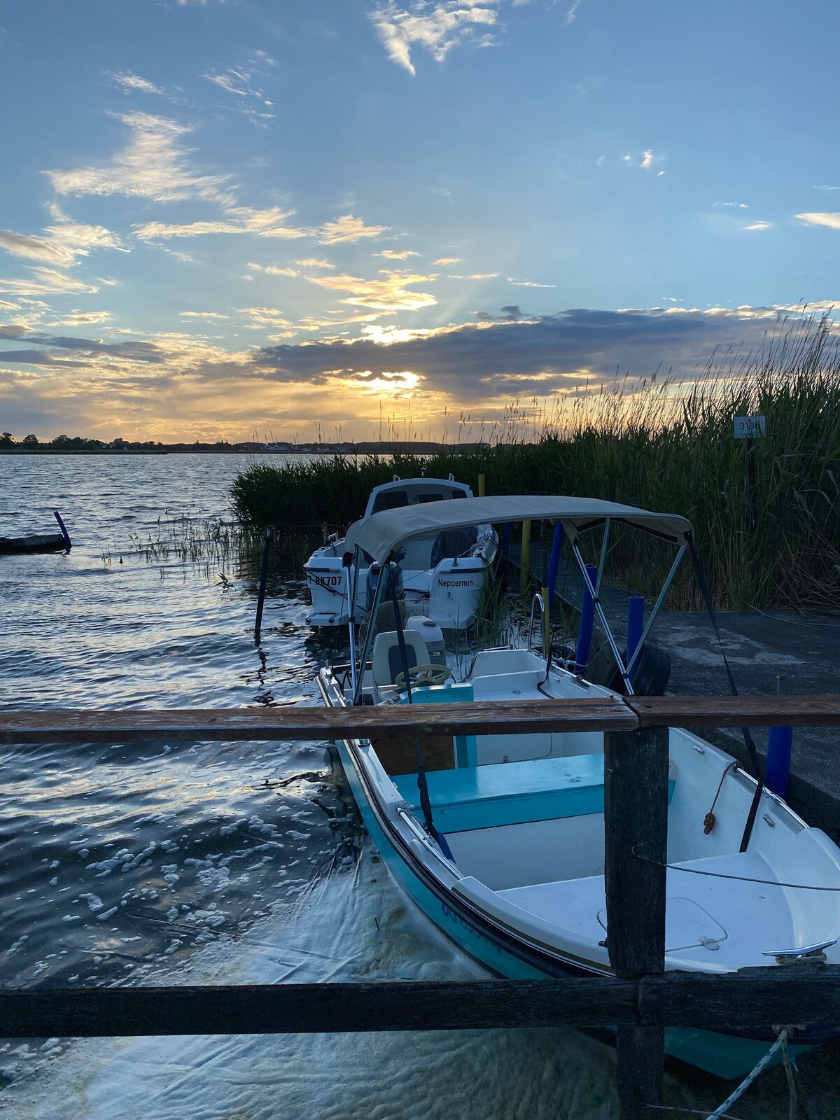 A boat is moored next to a wooden dock, with gentle ripples disturbing the water's surface. The sky is painted with soft hues of blue and orange as the sun sets, surrounded by tall reeds that frame the serene scene.