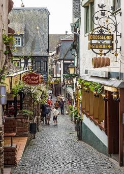 A cobblestone street lined with charming, historic buildings. Small shops and restaurants are adorned with decorative signage and greenery. People are seen strolling leisurely, creating a welcoming atmosphere. The street leads through a picturesque area, typical of a quaint village setting.