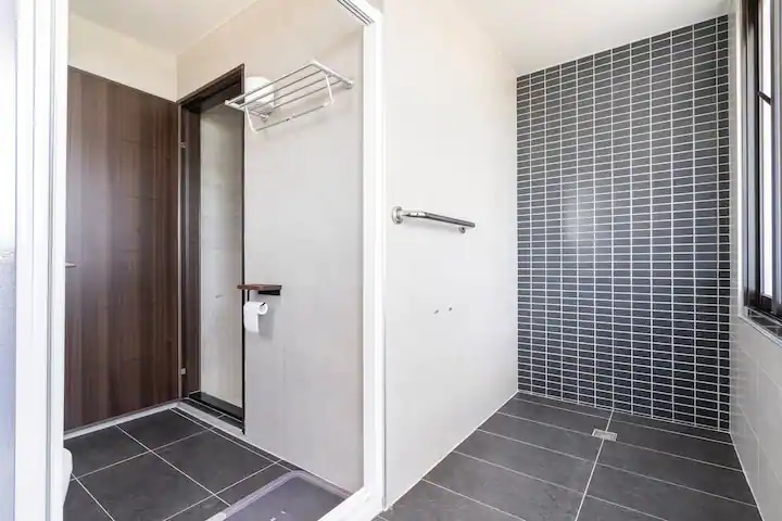 A modern bathroom is showcased, featuring sleek gray tiles on the floor and walls. A walk-in shower with glass doors is present, along with a towel rack and a toilet. Ample natural light is allowed through a window, enhancing the space's cleanliness.