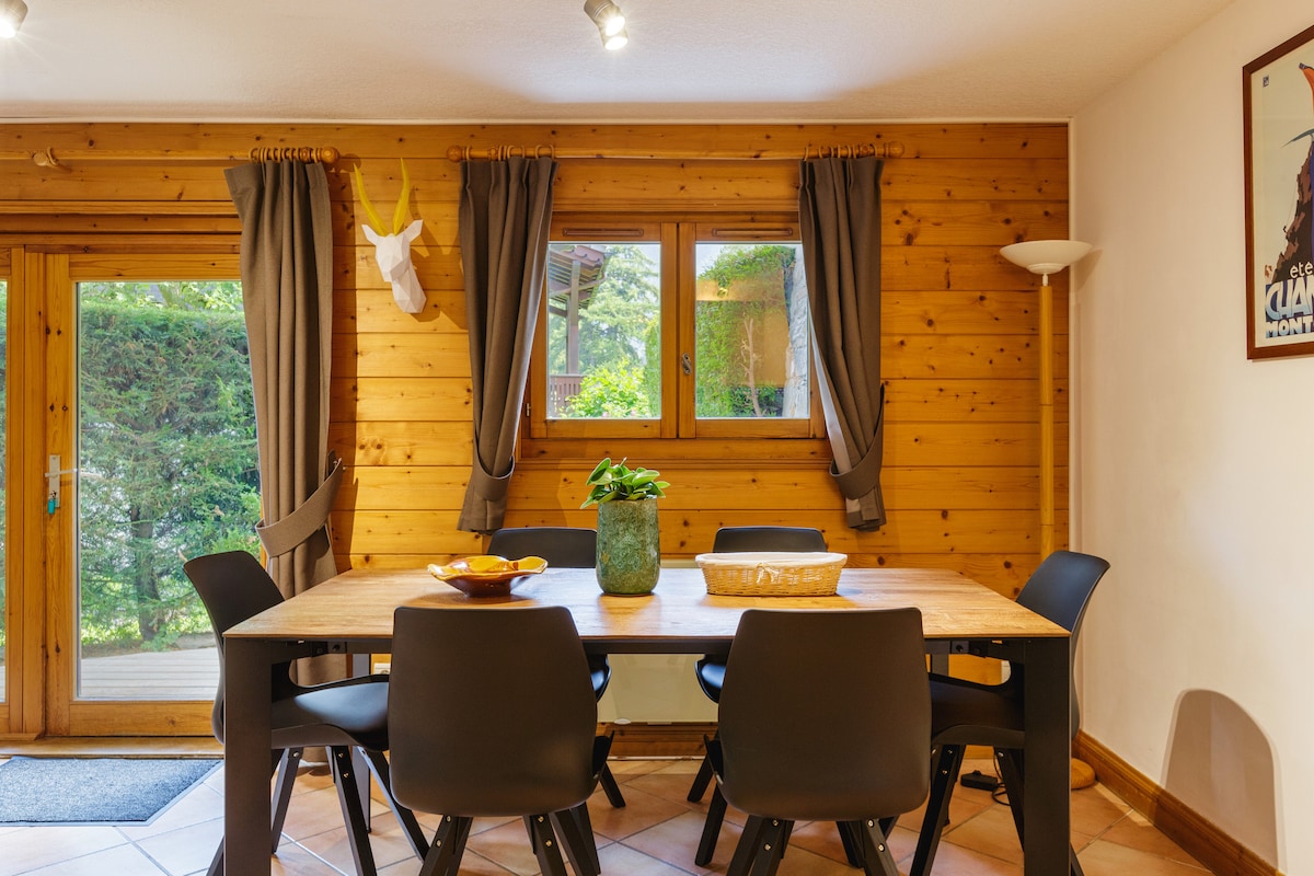 A dining area is showcased with a large wooden table surrounded by six black chairs. Natural light filters through a window adorned with grey curtains, highlighting wooden wall accents and a decorative deer head mounted above the table. A potted plant and bowls rest on the table.