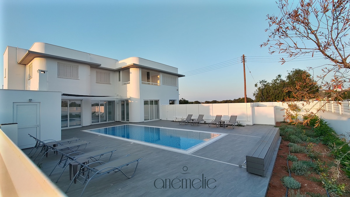 A modern, two-story holiday home is showcased with a spacious swimming pool in the foreground. Sun loungers are arranged around the pool, and a white fence provides privacy. The exterior features large windows, allowing natural light into the living spaces.
