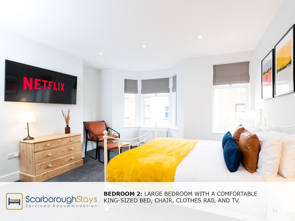 A spacious bedroom is presented with a comfortable king-sized bed adorned with soft white linens. A chair and a wooden dresser provide functional storage, while a clothes rail offers additional space. Natural light fills the room through large windows, complemented by a wall-mounted TV featuring Netflix.