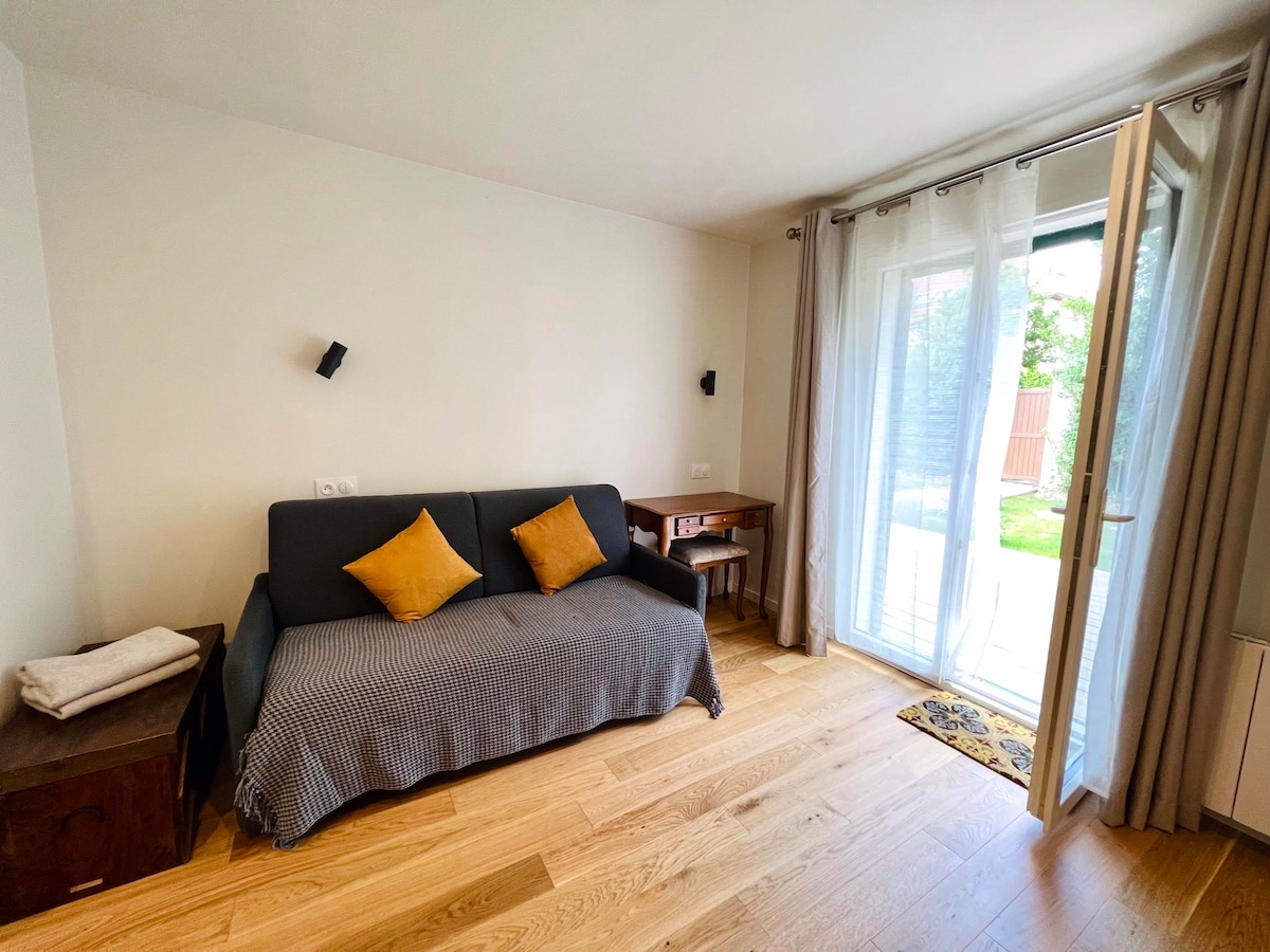 A bright living area features a convertible sofa adorned with cushions, positioned next to a wooden desk. Natural light floods in through a set of French doors that open to the private garden, enhancing the room's serene ambiance.