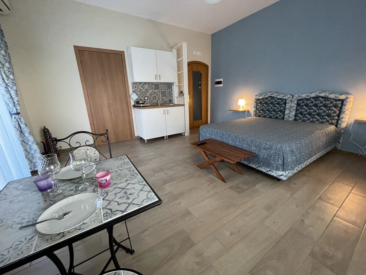 A spacious room features a comfortable bed with a patterned bedspread. A small dining table is set for two with plates and glasses, while a kitchenette is conveniently positioned in the corner. Natural light fills the space through a window dressed in light curtains.