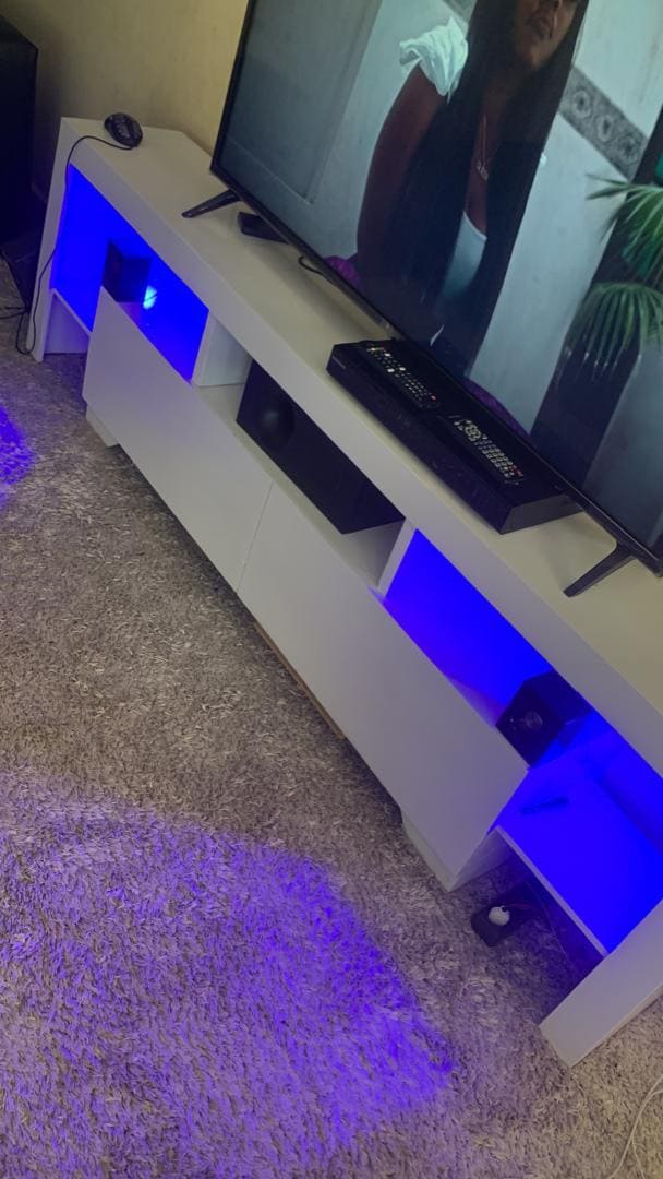 A modern entertainment unit is featured, illuminated by soft blue lights. The unit provides storage with open compartments, housing speakers and equipment. A large television is mounted above, displaying a scene from a film, while a plush carpet adds warmth to the area.