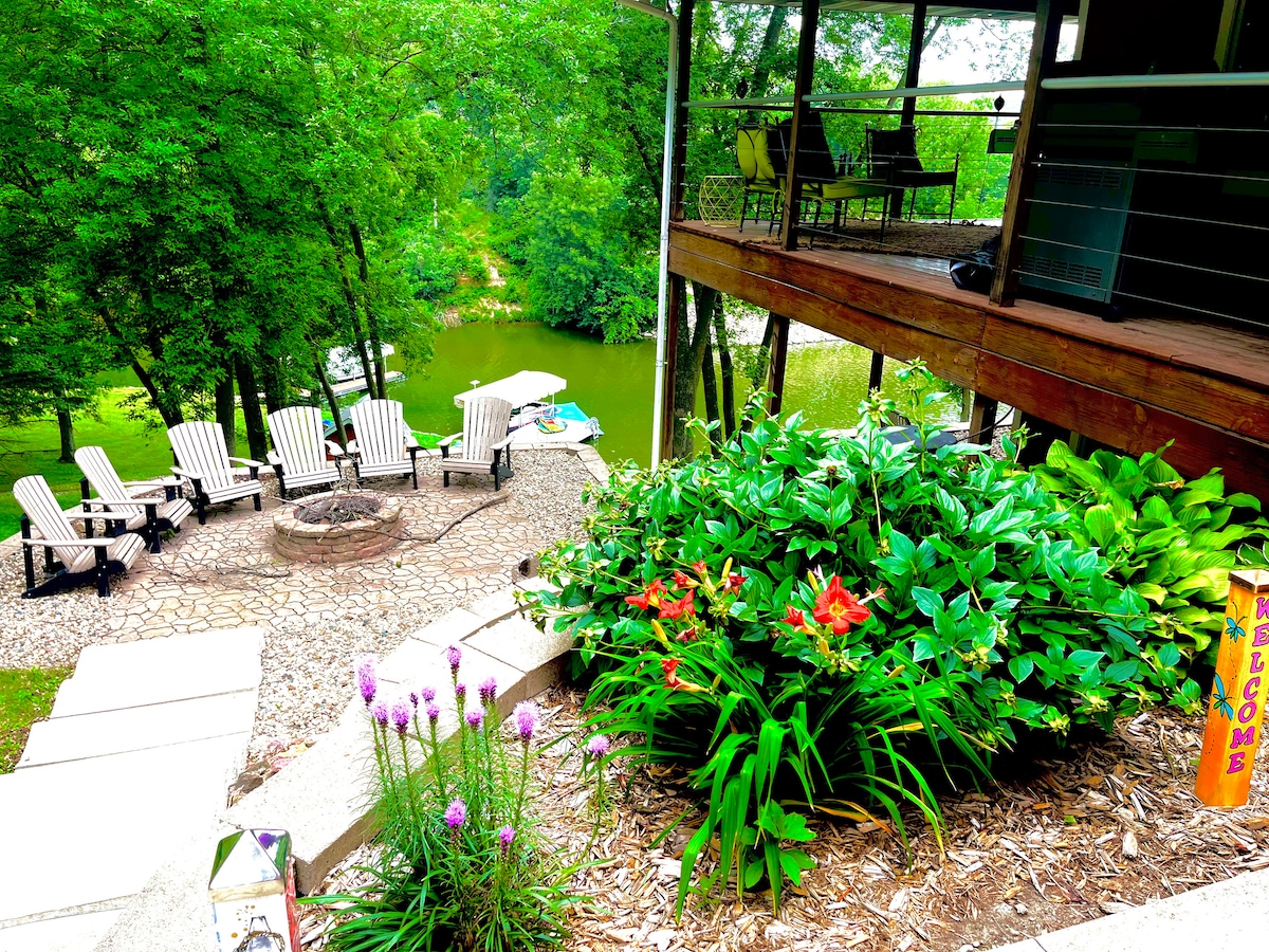 A tranquil outdoor area is presented, featuring a circular fire pit surrounded by several Adirondack chairs. Lush greenery and colorful flowers enhance the landscape, with steps leading down toward the lake in the background. A covered deck creates a shaded space above.