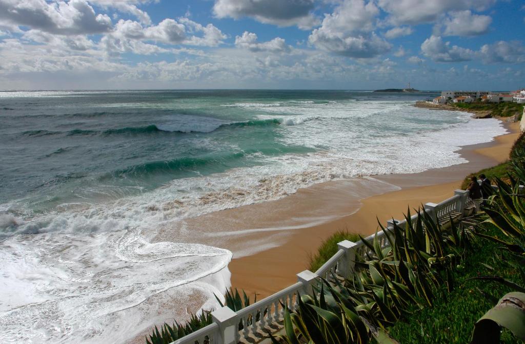 A scenic coastal view captures rolling waves crashing onto a sandy beach, bordered by lush greenery. The vibrant ocean reflects hues of blue and green, under a sky featuring scattered clouds, creating a serene atmosphere typical of Mediterranean landscapes.