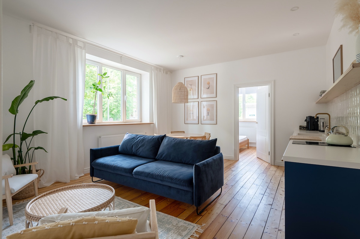 A spacious living area features original wooden flooring and high ceilings. A large blue sofa is positioned in front of a window with natural light illuminating the space. A dining area for four and a well-equipped kitchen are visible in the background.