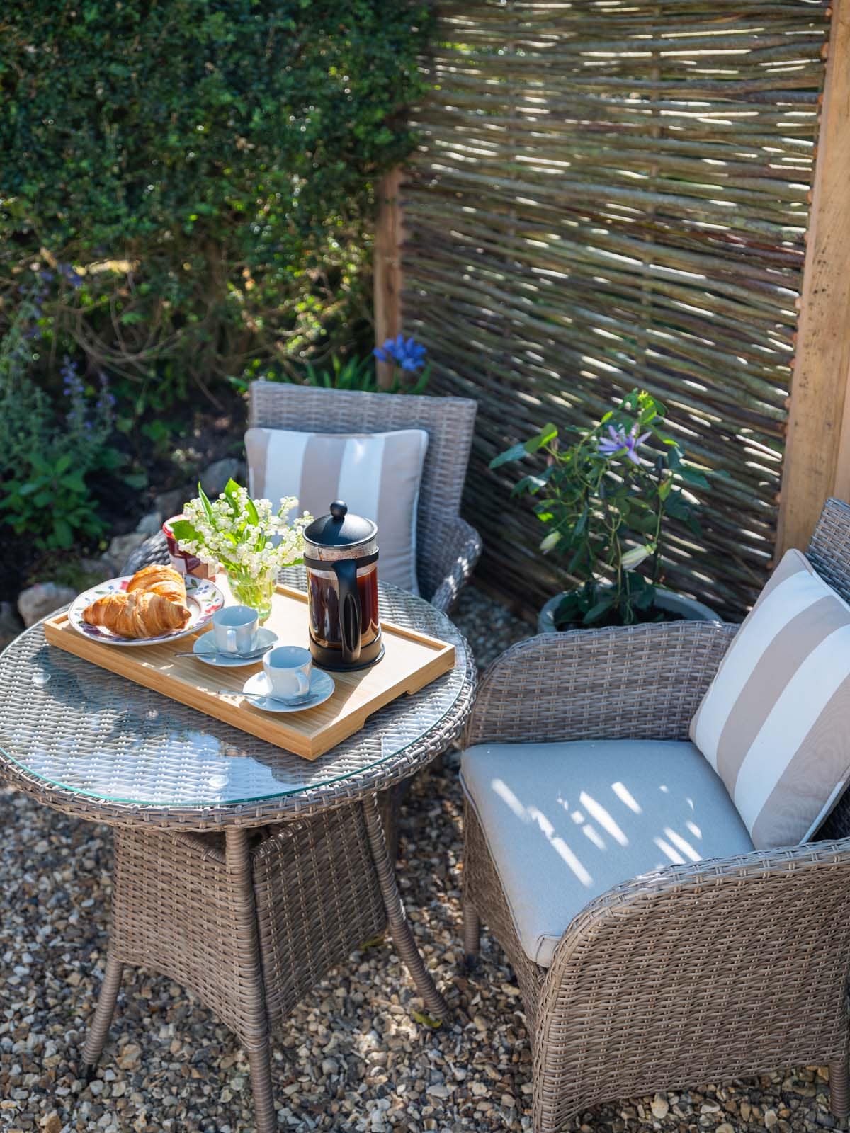 A quaint outdoor seating area is depicted with two woven chairs and a round table topped with a tray. The tray holds a French press, coffee cups, and freshly baked pastries. Sunlight filters through leaves, casting gentle shadows on the gravel surface.