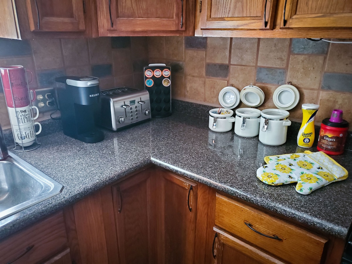 A fully equipped kitchen countertop is featured, showcasing a coffee maker, toaster, and a set of canisters. Kitchen towels and patterned oven mitts are positioned nearby. A small sink is visible, surrounded by wooden cabinetry and decorative tile backsplash.