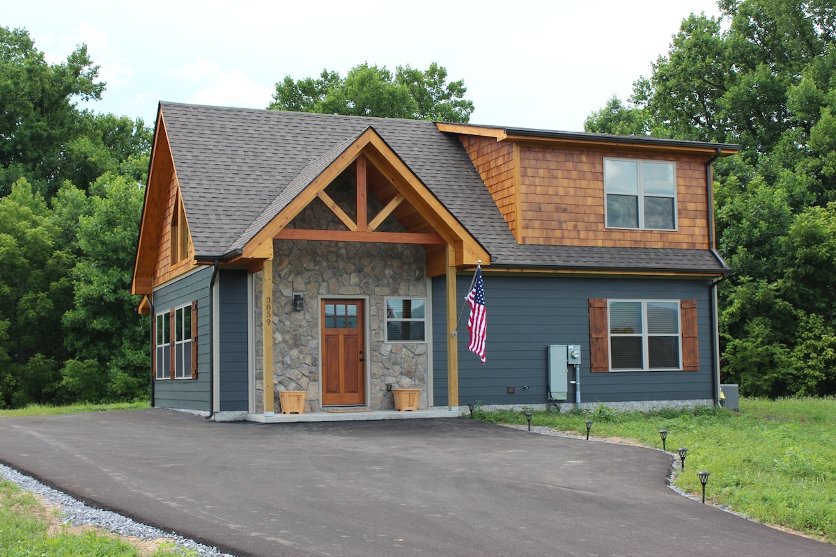 A newly constructed cabin features a blend of stone and wood siding, accentuated by a sloping roof. A welcoming front door with large windows is framed by two planters. An American flag hangs proudly beside the entrance, enhancing the cabin's charming exterior.