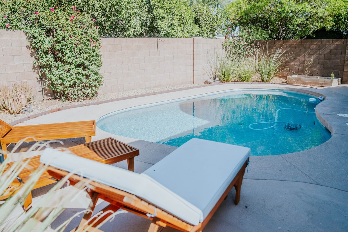 A private backyard is depicted featuring a pool surrounded by a concrete patio. Sun loungers are positioned near the pool, providing a relaxing space. Lush greenery and a stone wall create a serene, enclosed atmosphere.