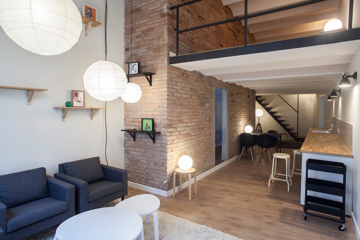 An open-plan space features a living area with two armchairs and a round coffee table. A light-colored rug adds warmth to the wood flooring. Soft lighting is provided by paper lanterns and wall-mounted fixtures. A set of stairs leads to additional living space above.