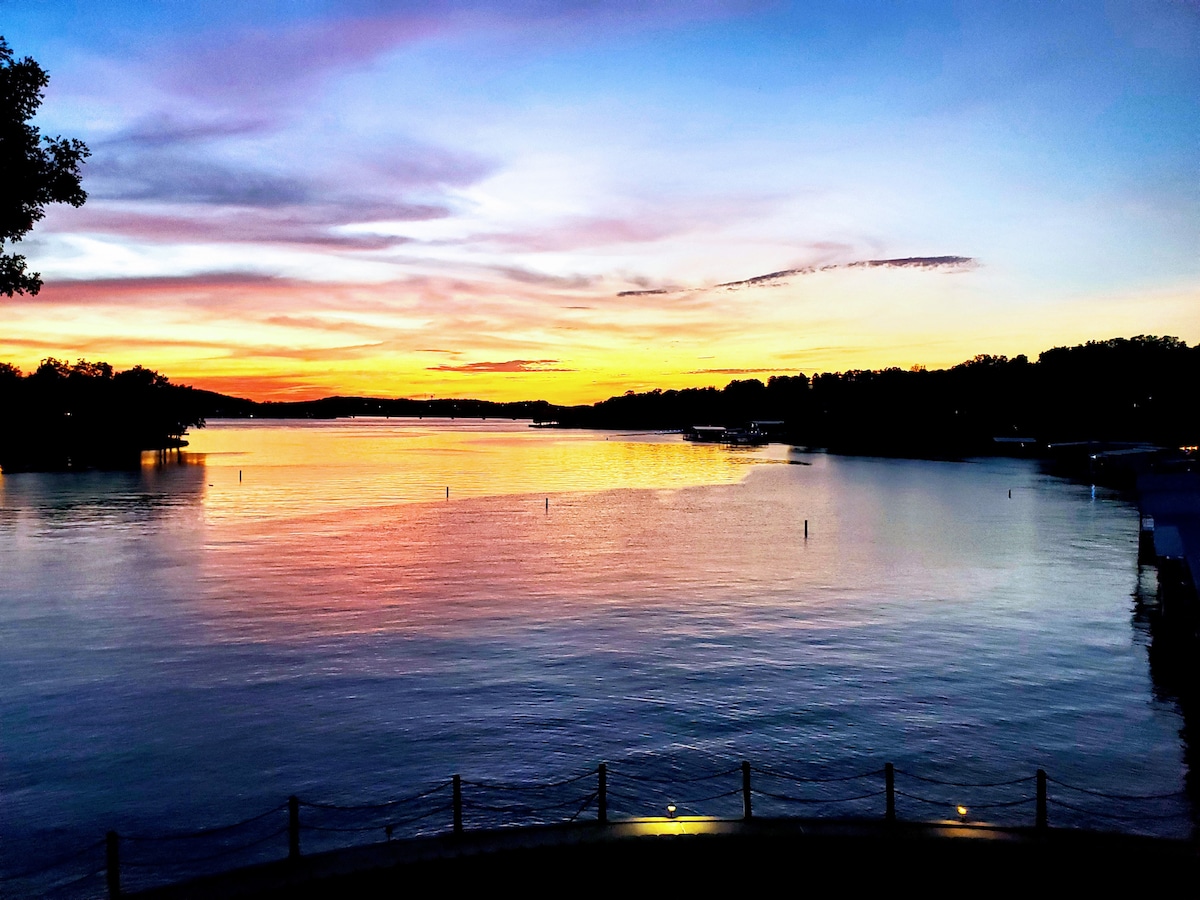 A stunning lake view at sunset showcases vibrant colors reflecting off the water. The horizon features a gradient of orange, pink, and purple hues, while silhouettes of trees line the shore, creating a serene atmosphere.