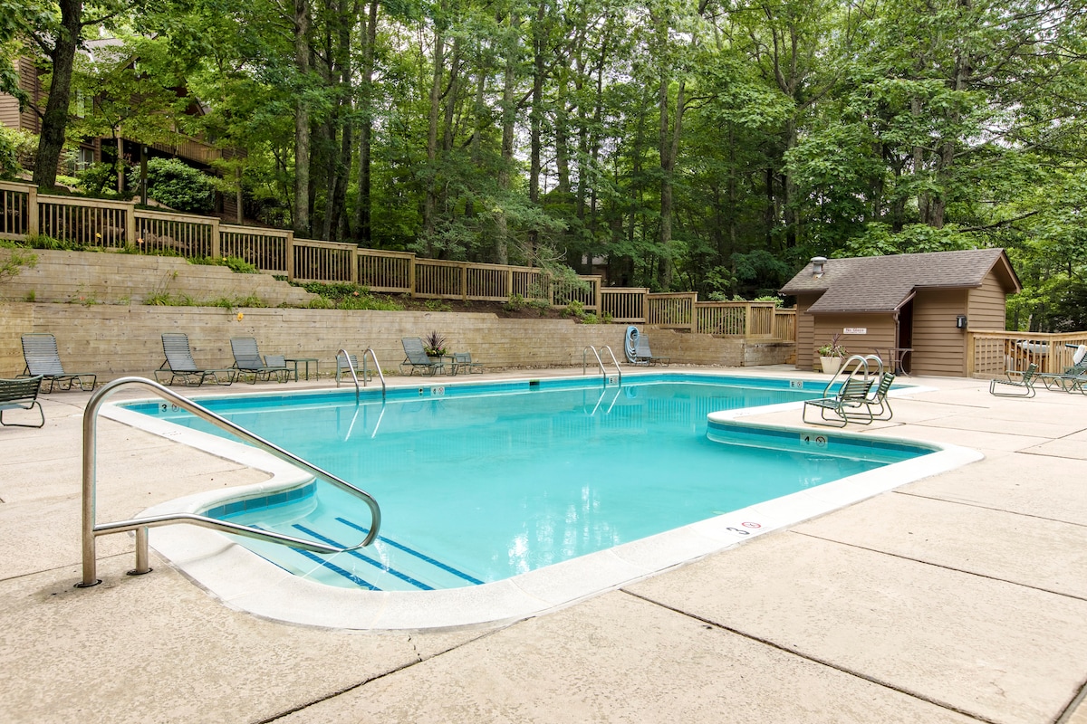 A shared outdoor pool is surrounded by a concrete patio with several lounge chairs. Lush greenery from trees provides a natural backdrop, and a wooden fence encloses the area. A storage shed is visible nearby, contributing to the functional space.