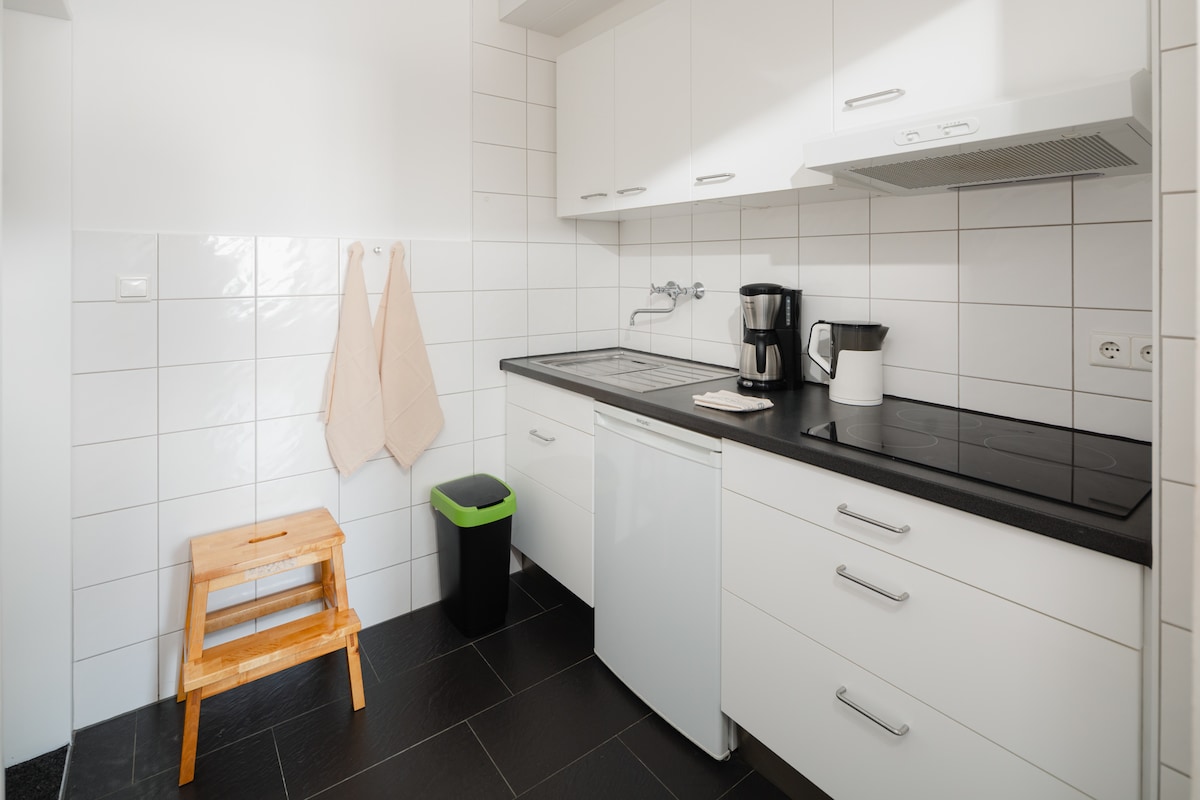 A functional kitchen area features a compact design with white cabinetry and a black countertop. Essential appliances, including a coffee maker and kettle, are visible. Natural light reflects off the tiled walls, while a wooden stool and a green trash bin provide additional utility.