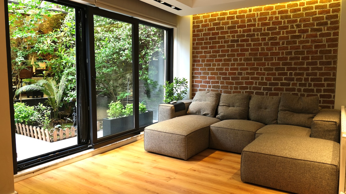 A cozy living area features a sectional sofa in textured fabric, positioned against a warm brick wall. Large glass doors provide a view of lush greenery, allowing natural light to fill the space. Warm wood flooring adds to the inviting ambiance.