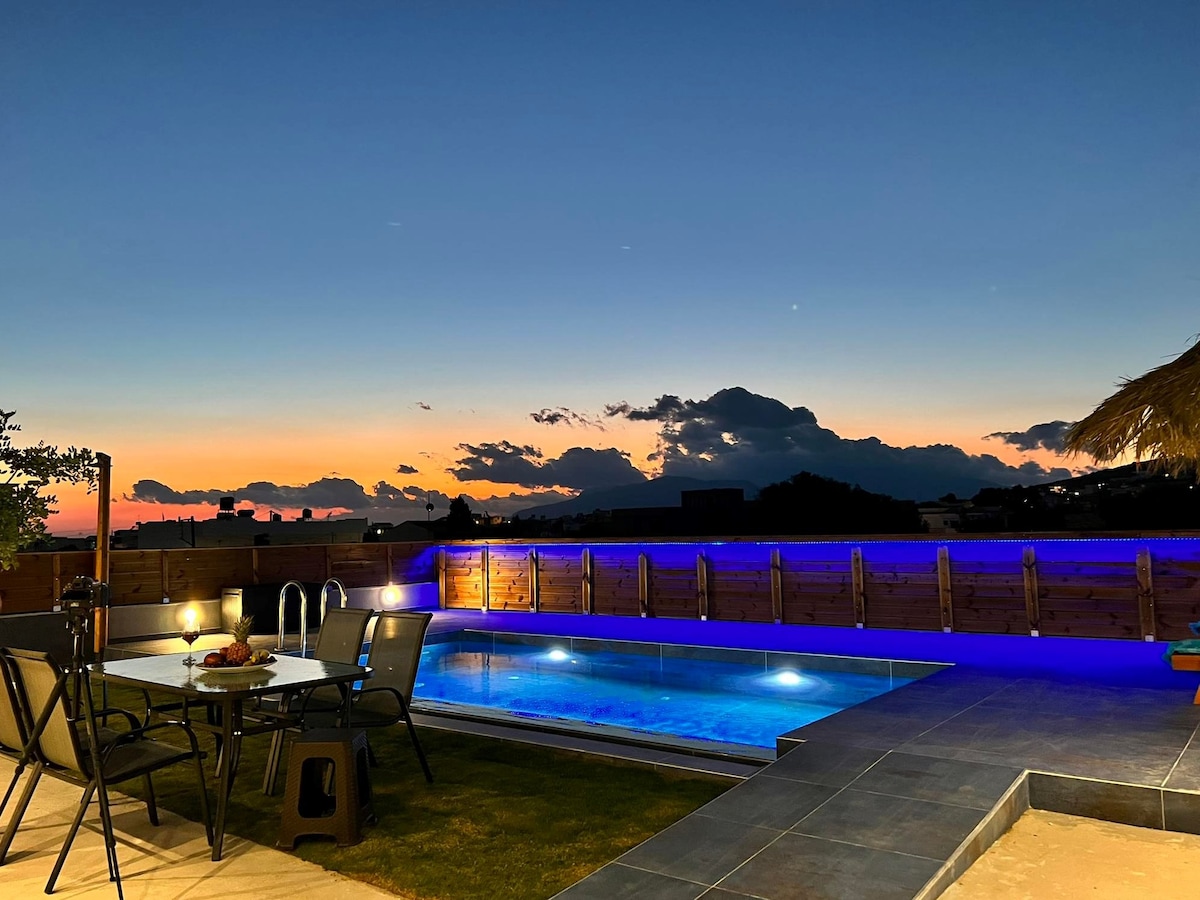An evening view captures a tranquil outdoor space featuring a pool illuminated with soft blue lighting. A dining table with chairs is positioned nearby, surrounded by lush greenery and bathed in the warm hues of sunset clouds.