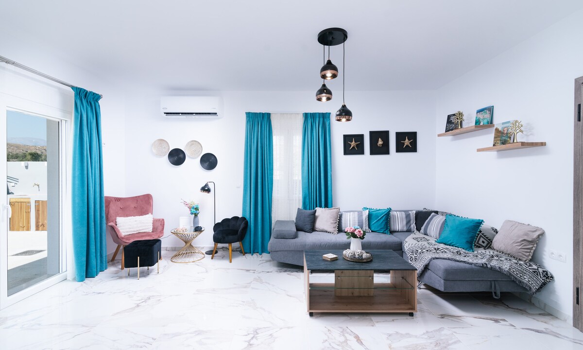 A modern living area features a spacious gray sectional sofa and a pink accent chair. Decorative elements include a wooden coffee table and two small side tables. Large windows adorned with turquoise curtains allow natural light to fill the room, enhancing the contemporary decor.