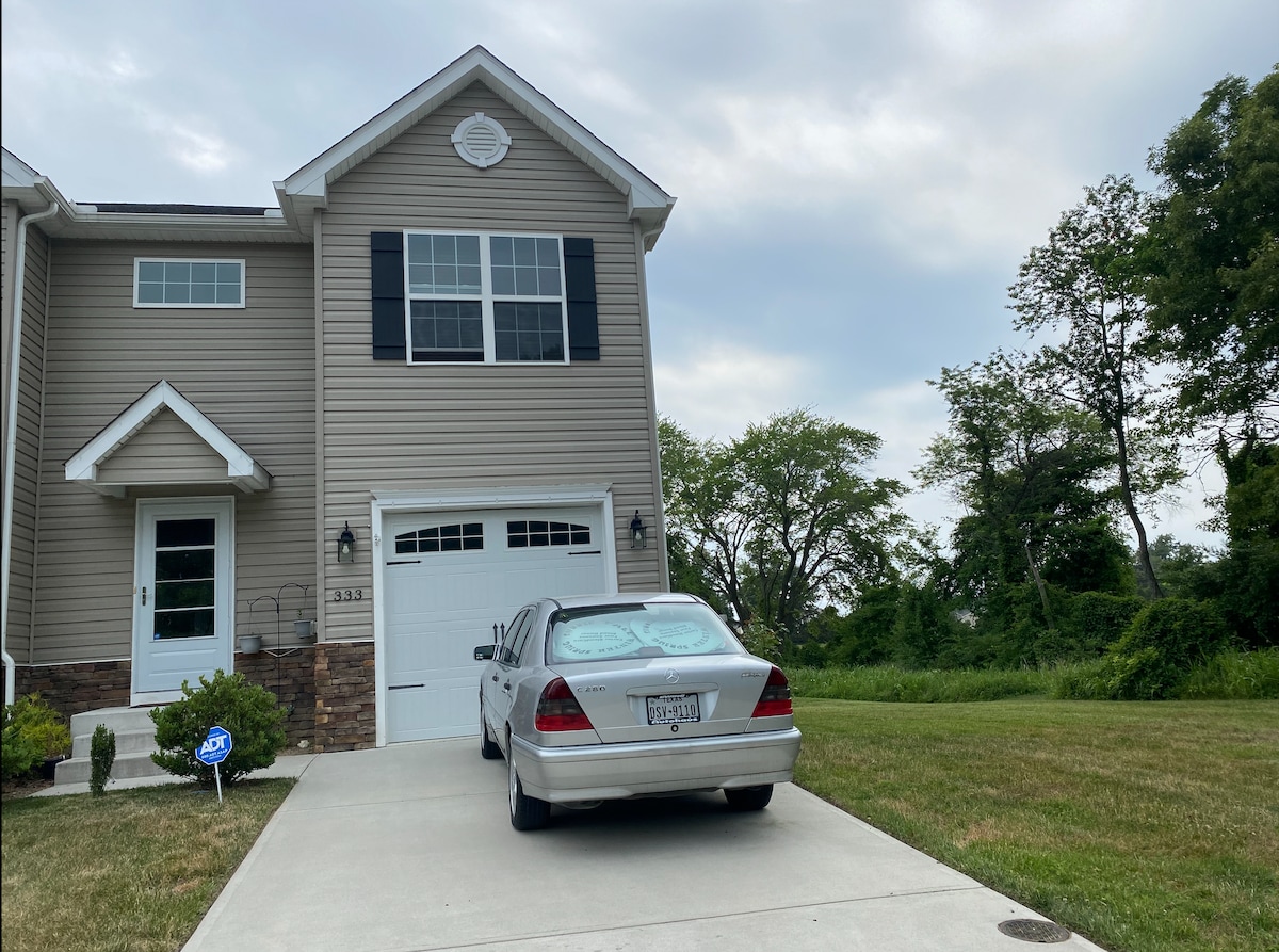 A townhome exterior is depicted, featuring a light brown facade with blue shutters and a covered entryway. A silver car is parked in the driveway, which leads to a manicured lawn and green shrubs. A grassy area extends in the background.