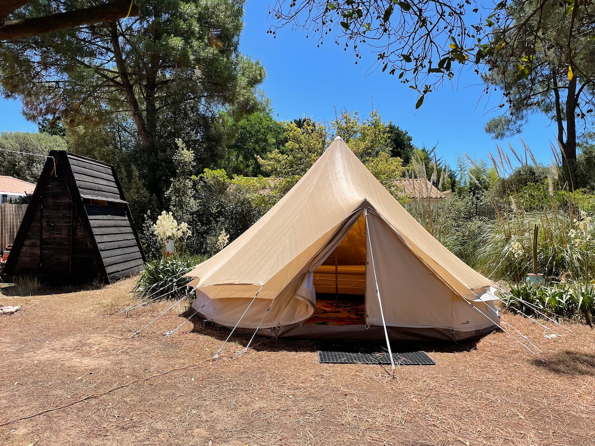 A spacious beige tipi tent is set in a natural garden surrounded by greenery and trees. The entrance is open, revealing a cozy interior. Nearby, a wooden structure and various plants enhance the tranquil outdoor setting.