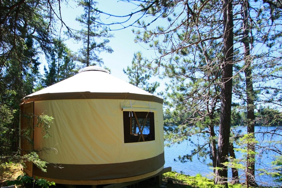A spacious yurt stands amongst tall pine trees, overlooking calm lake waters. Large windows provide views of the serene surroundings, while the tent-like structure blends harmoniously with nature.