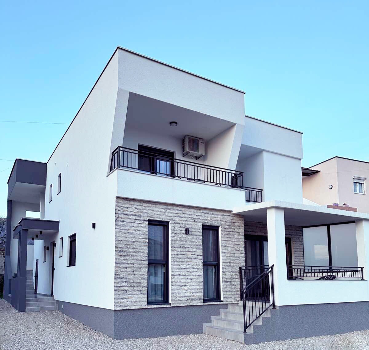 A modern two-story house features a combination of smooth white walls and textured stone accents. Large windows allow natural light to fill the interior. A covered balcony is visible, providing an outdoor space that overlooks the surroundings. A simple staircase leads to the entrance.