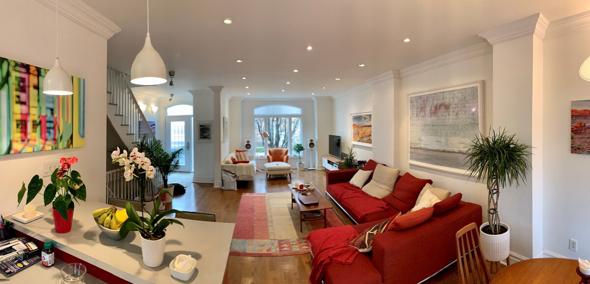 The open-concept living area features bright, recessed lighting and modern furnishings. A red sectional sofa and armchairs provide ample seating. A coffee table is placed centrally on a patterned area rug, and large windows allow natural light to flow in, enhancing the spacious atmosphere.