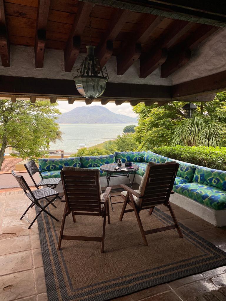 A covered outdoor seating area features a circular table surrounded by wooden chairs and a colorful bench. Lush greenery and the lake view are visible in the background, creating a serene setting. Natural light filters through, enhancing the inviting ambiance.