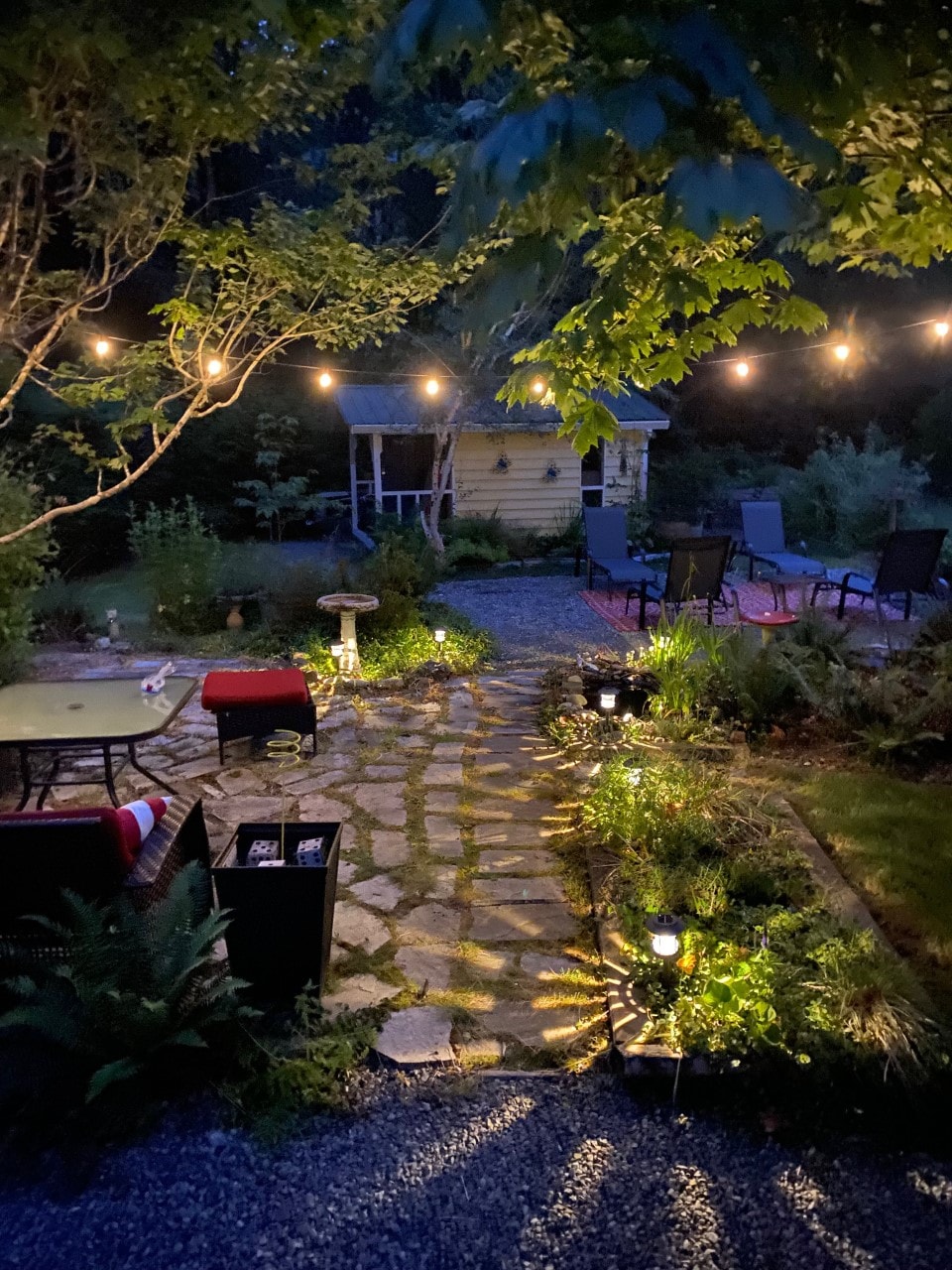 The evening garden is illuminated by string lights, casting a warm glow over a stone path. A seating area with chairs and table is visible, surrounded by lush greenery and soft lighting from solar lamps, creating a serene outdoor space.