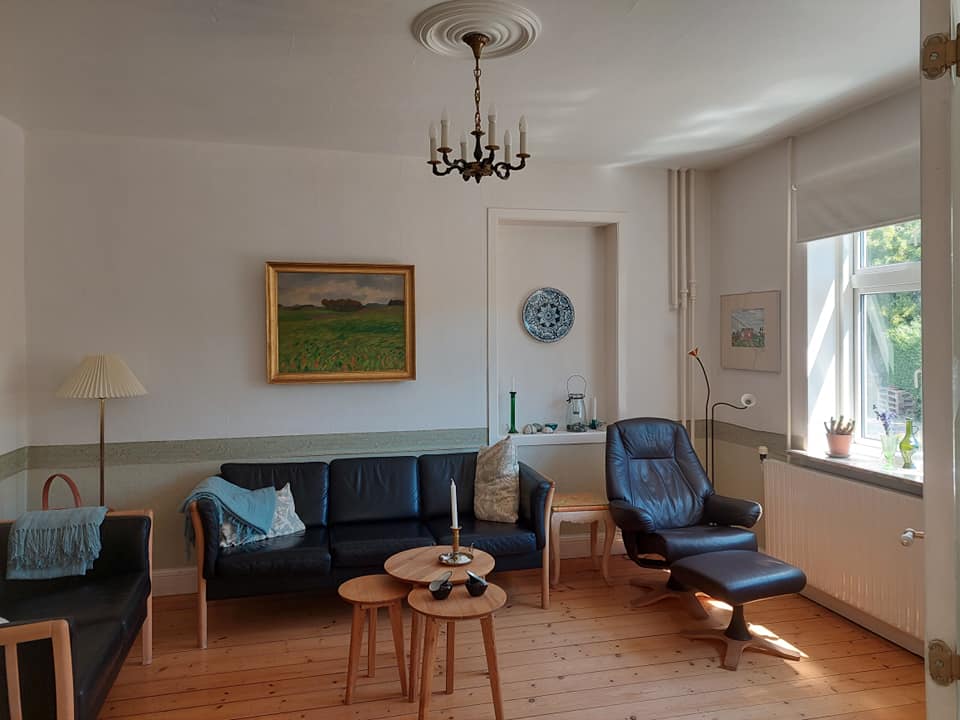A cozy living area features a combination of comfortable seating, including a dark leather armchair and a sectional sofa. A pair of wooden side tables is positioned at the center, and natural light enters through a large window adorned with light curtains. Decorative elements include a chandelier and artwork on the walls.
