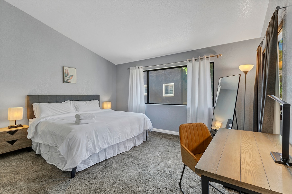 The bedroom is spacious and features a large bed with white linens. Soft light is provided by two bedside lamps. A window is dressed with sheer curtains, allowing natural light to brighten the room. A wooden desk and chair are positioned near the window, alongside a full-length mirror.