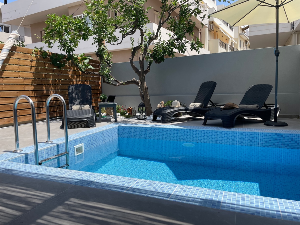 A private pool is framed by lounge chairs and a shade umbrella, surrounded by a low wall. Lush greenery is visible, adding natural elements to the outdoor space. The clear water reflects sunlight, creating a serene environment for relaxation.