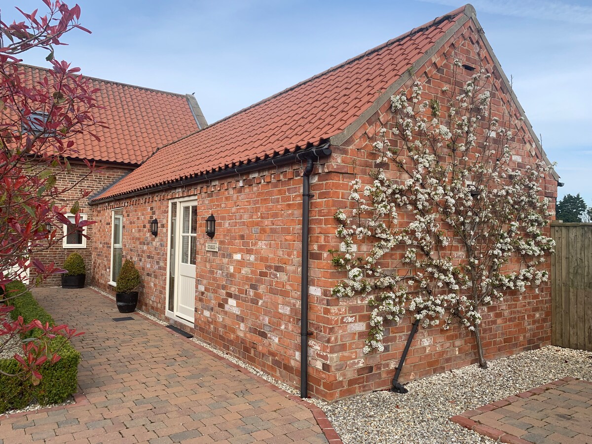 A charming barn exterior, featuring a red brick façade and a sloped roof with terracotta tiles. Flowering plants are visible along one wall, contributing to the serene environment. The paved pathway leads to the entrance, surrounded by neatly trimmed bushes.