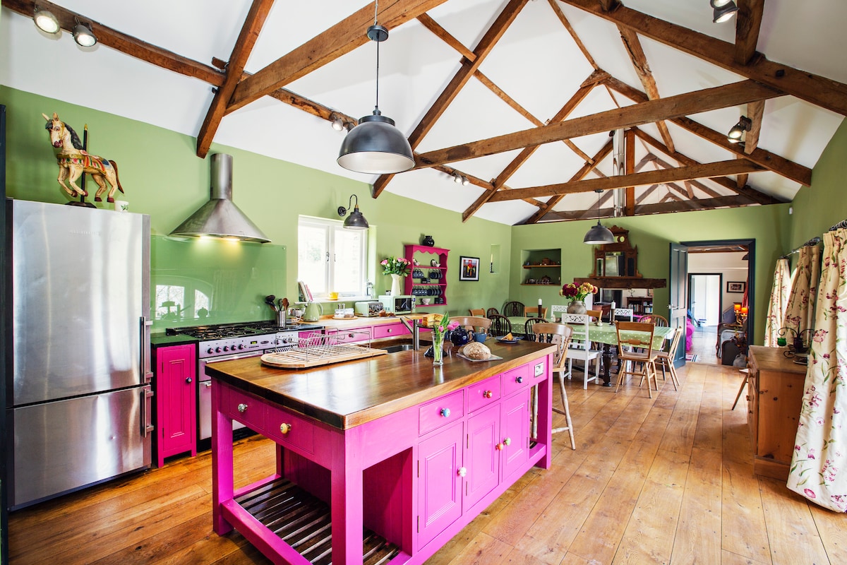 A vibrant kitchen features a large pink island with a wooden countertop, complemented by stainless steel appliances. High ceilings with exposed wooden beams create openness, while green walls add warmth. A dining area with a rustic table and chairs is visible in the background, contributing to the inviting atmosphere.
