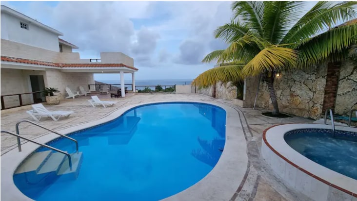 An inviting outdoor area features a large pool surrounded by a spacious deck with lounge chairs. A jacuzzi is present nearby, and palm trees provide shade. The ocean can be seen in the distance, creating a relaxed ambiance.