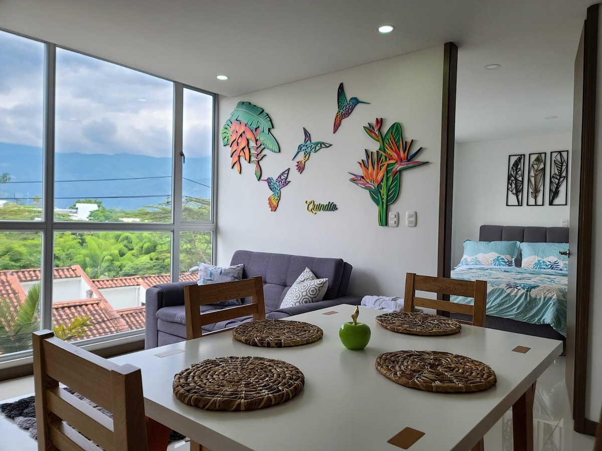 A modern living space is shown, featuring a dining table with woven placemats and a decorative centerpiece. A comfortable sofa is positioned against a large window, which offers a view of greenery and mountains. Colorful tropical wall art enhances the ambiance.