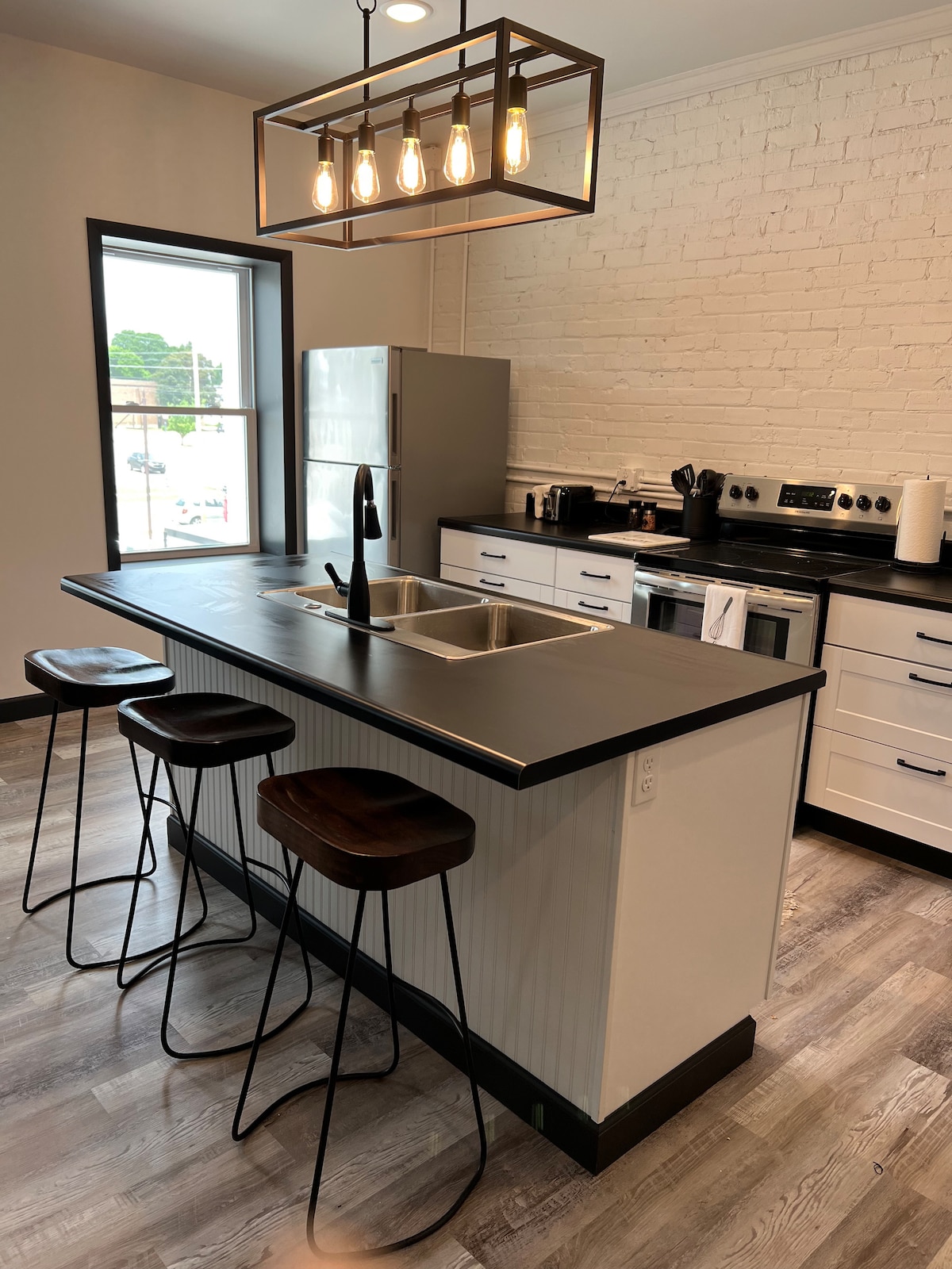 The modern kitchen features a sleek black island with seating for three. Stainless steel appliances and white cabinetry complement the brick wall. A pendant light hangs above the island, illuminating the workspace.