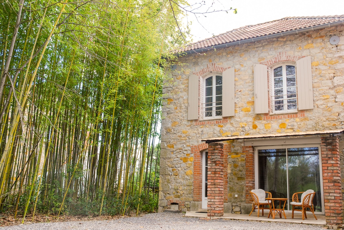 The exterior of the stone house features large windows with shutters, framed by lush bamboo greenery. A patio area is visible, showing two wicker chairs situated on gravel, providing a peaceful outdoor space. The rustic structure is complemented by a brick column design.