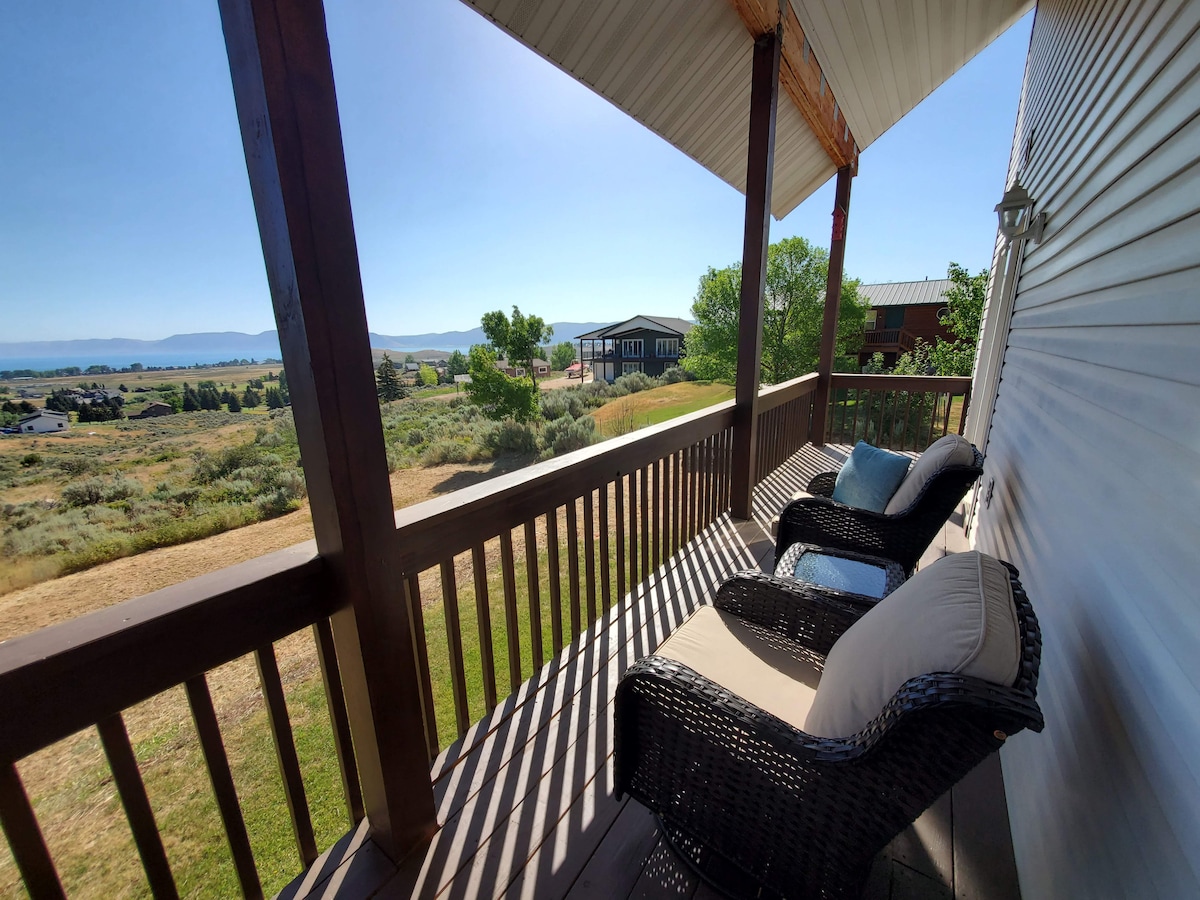 A serene wraparound deck is featured, furnished with two comfortable wicker chairs. The view captures a clear sky and expansive greenery, leading to water in the distance. Sunlight brightens the outdoor space, inviting relaxation in a tranquil setting.