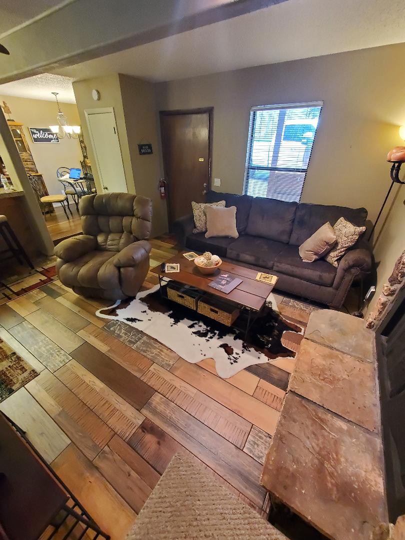 A comfortable living area showcases a spacious sectional sofa paired with a recliner. A wooden coffee table, adorned with decorative items, rests on a patterned rug. Natural light filters through the nearby window, illuminating the warm wood flooring and the cozy ambiance of the space.