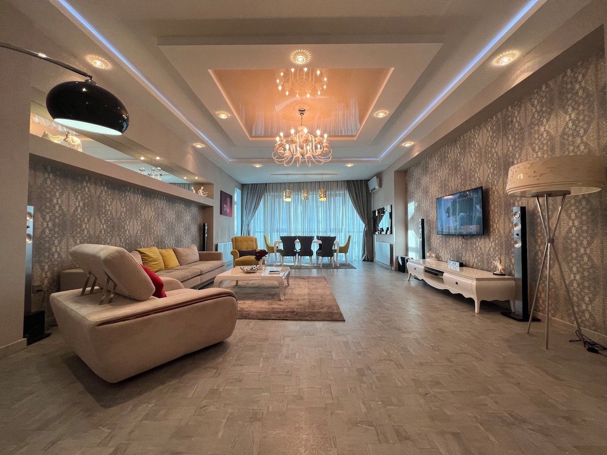 A spacious living area features a modern sofa arrangement with colorful cushions. A dining table with multiple chairs is positioned near large windows that allow natural light. An elegant chandelier hangs from the ceiling, complementing the contemporary decor and patterned wallpaper.