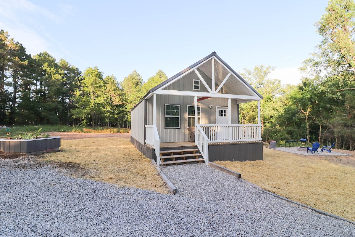 A newly constructed cabin is surrounded by a gravel pathway and grass, featuring a front porch with stairs leading to the entrance. The exterior showcases light-colored siding and a gabled roof, with large windows that provide natural light to the interior.
