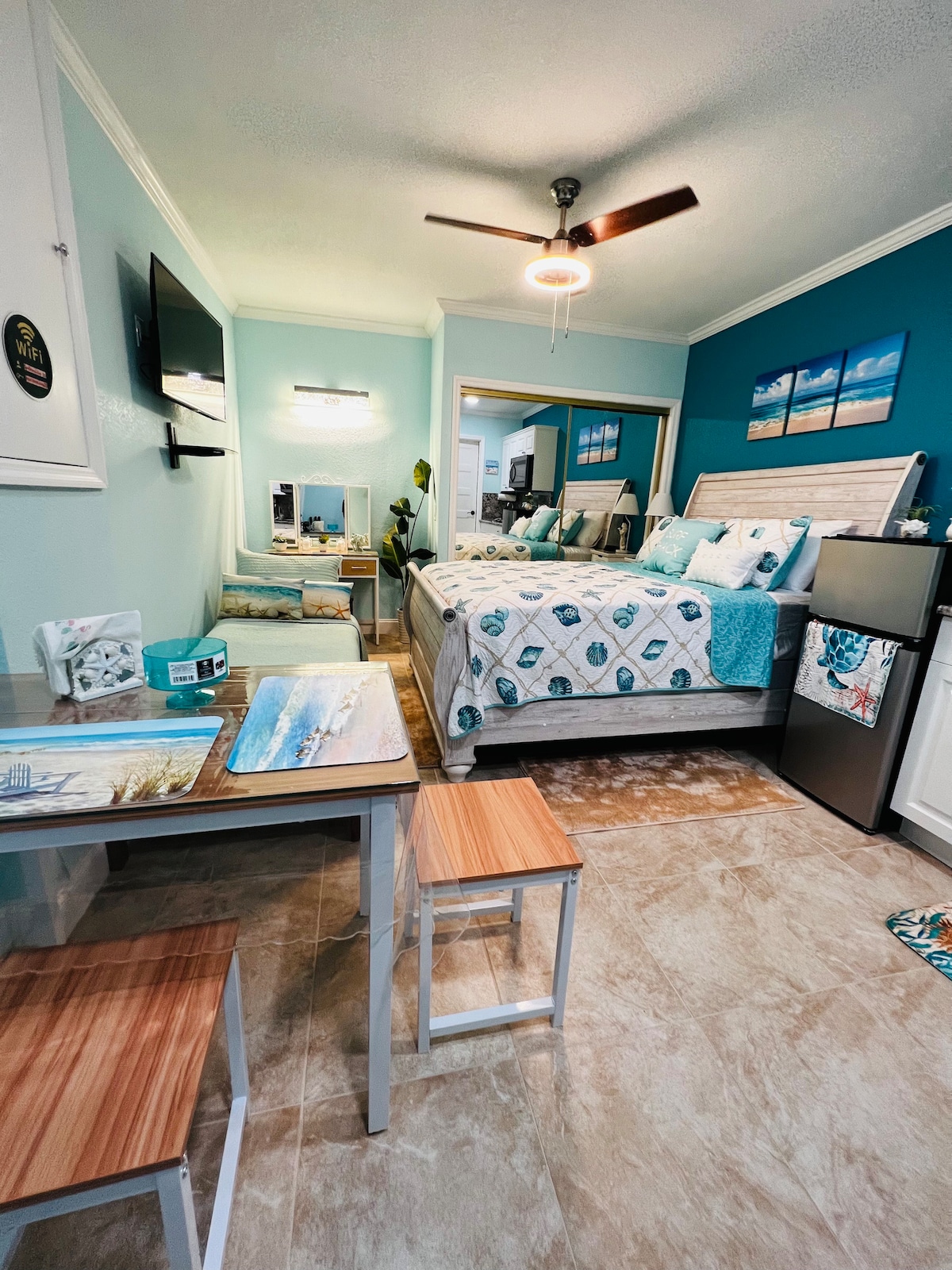 A cozy and modern room features a spacious bed with blue and white bedding set against an accent wall. A small table with two stools is situated nearby, and a refrigerator is visible in the corner. Natural light brightens the area, complemented by decorative beach-themed artwork.