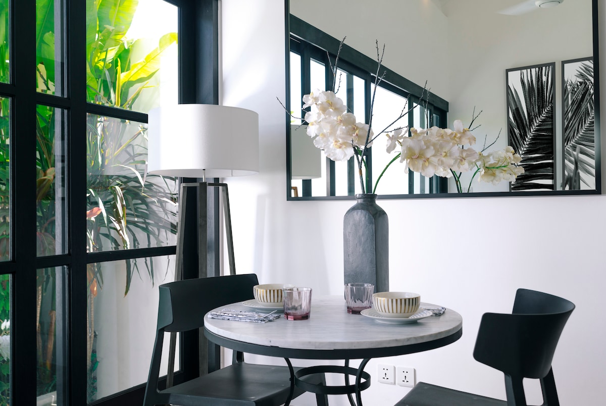 A modern dining area is presented with a round marble-topped table and two black chairs, complemented by decorative tableware. A tall vase with white flowers adds elegance, while large windows allow natural light and showcase lush greenery just outside.