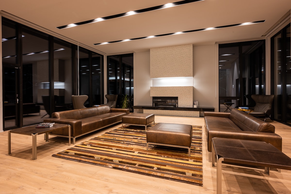 The spacious living area is designed with sleek leather sofas arranged around a central coffee table. A modern fireplace is featured on the wall, surrounded by large windows that allow natural light to fill the room.