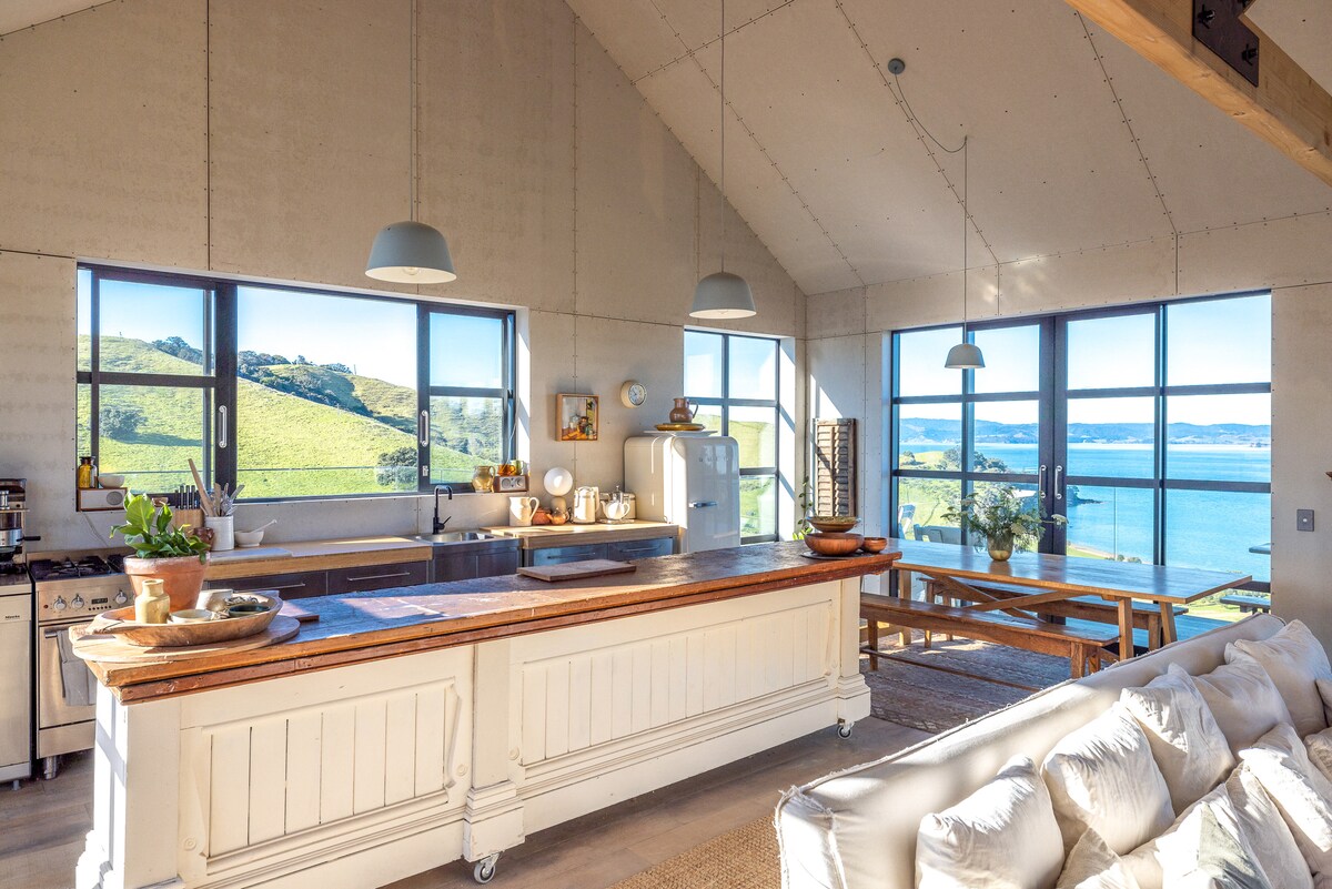An open plan kitchen and dining area features large windows providing panoramic views of the surrounding landscape and water. A spacious wooden island and rustic dining table are complemented by modern lighting. Natural light floods the space, highlighting the contemporary industrial design.