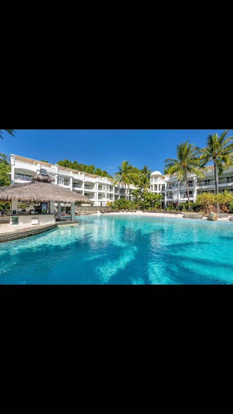 The resort features a large swimming pool surrounded by palm trees and lush greenery. A swim-up bar is visible under a thatched roof, offering a shaded area for relaxation. The modern building stands in the background, complemented by bright blue skies.