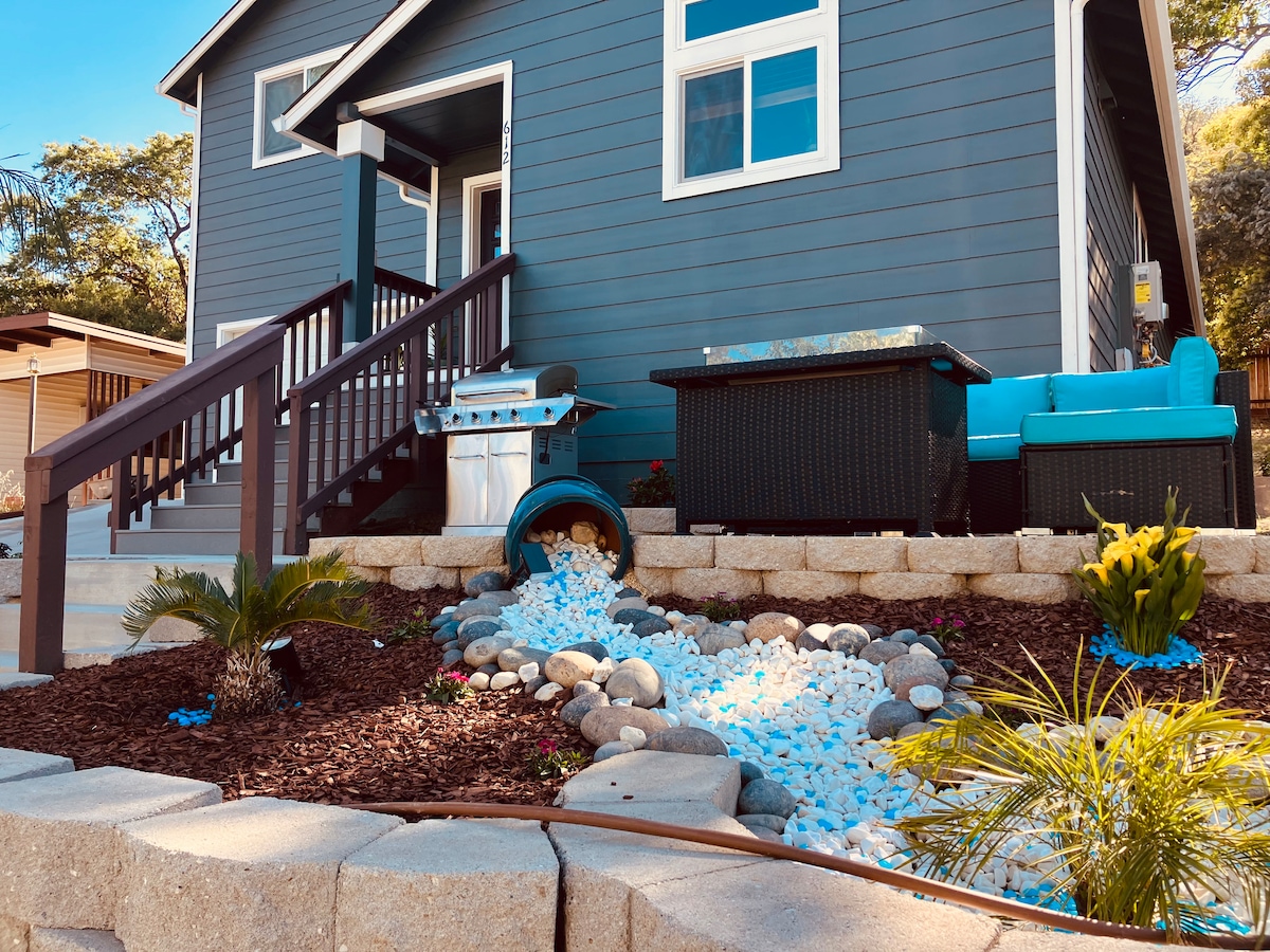 A modern home is presented with a well-maintained landscape featuring a decorative rock garden. A shallow water feature runs through smooth stones, surrounded by vibrant greenery. Comfortable seating is arranged nearby, complimented by a barbecue grill positioned on a wooden deck with steps leading to the entrance.